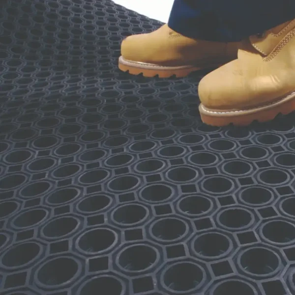 A person wearing yellow work boots stands on a Roof Walkway Mat featuring a black rubber surface with a circular hole pattern.