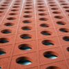 Close-up of the Food Production Mat’s textured grid of reddish-brown tiles, each featuring a central circular hole, creating a repetitive geometric pattern illuminated from above.