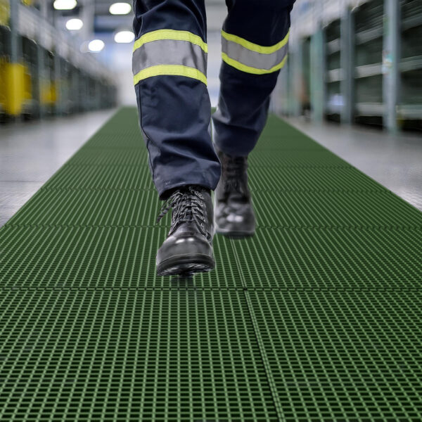 Close-up of a person wearing black work boots and navy trousers with reflective stripes, walking on Open Top Grating in an industrial setting. Shelving and bright lighting are visible in the background.