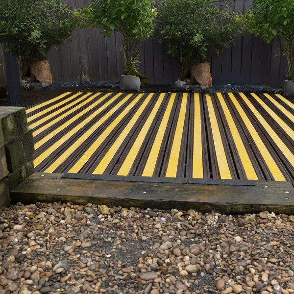 Anti-Slip Decking Strips in yellow and black are laid between wooden planks, bordered by potted trees along a dark fence, with pebbles in the foreground.