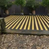 A garden deck features Anti-Slip Decking Strips in alternating yellow and dark brown, bordered by potted trees along a dark fence, with a pebble path in front.