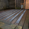 Wet and weathered wooden patio decking features several Anti-Slip Decking Strips attached in parallel lines, positioned beside a brick wall, a wooden fence, and a metal bin.