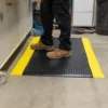 A person in tan work boots and black trousers stands on the Premium Bubble Top Anti-Fatigue Mat, which features a black and yellow design, in a workshop with various tools and equipment in the background.