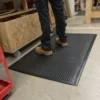 A person wearing black trousers and tan work boots stands on a Premium Bubble Top Anti-Fatigue Mat in a workshop, with wood and tools in the background.