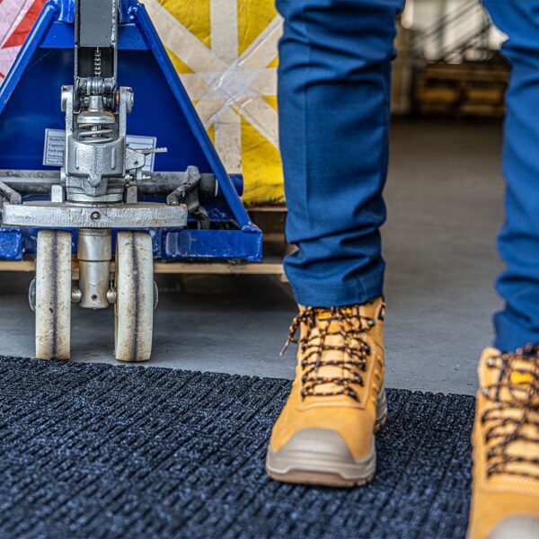 A person in blue trousers and tan work boots stands next to the wheel of a blue pallet truck on a Forklift Mat in an industrial setting.