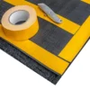 A roll of yellow double-sided tape, strips applied to a black surface, and a silver utility knife are arranged on a Forklift Mat.