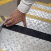 A hand applies Anti-Slip Comfortable Tape to a chequer plate metal stair tread, improving safety by adding a black anti-slip surface.