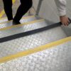 A person applies Anti-Slip Comfortable Tape, a black strip with yellow safety markings, to the edge of a metal stairway.