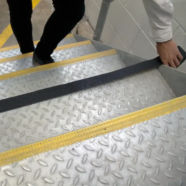 A person applies Anti-Slip Comfortable Tape, a black non-slip strip, to the edge of a metal stair with yellow safety lines in an industrial setting.