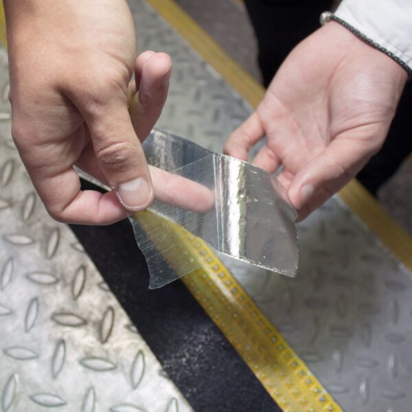 A person holds a piece of Anti-Slip Comfortable Tape above metal flooring featuring textured patterns and a yellow strip along the edge.