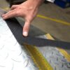 A person applies Anti-Slip Comfortable Tape, a black strip with a nearby yellow safety stripe, to a textured metal surface—possibly a stair tread—for enhanced grip.