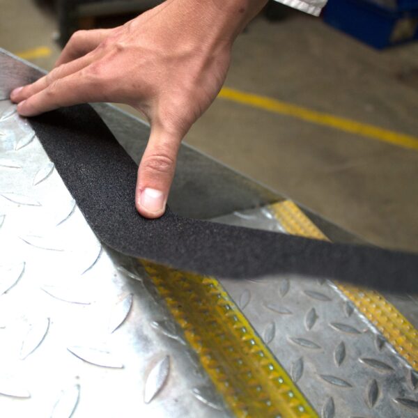 A person applies Anti-Slip Comfortable Tape, a black strip with a nearby yellow safety stripe, to a textured metal surface—possibly a stair tread—for enhanced grip.
