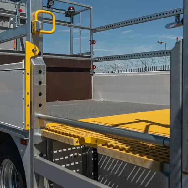 Close-up of a lorry featuring safety rails, a yellow handrail, and an Anti-Slip Flat Sheet platform. The lorry is parked near a barbed-wire metal fence under a blue sky.