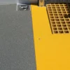 Close-up of a floor featuring the Anti-Slip Flat Sheet: textured grey surface on the left, smooth yellow centre, and a yellow grid-patterned mat on the right. A metal fixture is embedded in the top left corner.