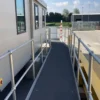 An Anti-Slip Flat Sheet is installed on a wheelchair-accessible ramp with metal railings, leading to a building entrance outdoors near a car park and industrial building, with green trees in the background.