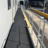 A narrow walkway featuring an Anti-Slip Flat Sheet with a black non-slip surface runs beside a building, bordered by metal railings. A construction vehicle and industrial equipment are visible in the background.