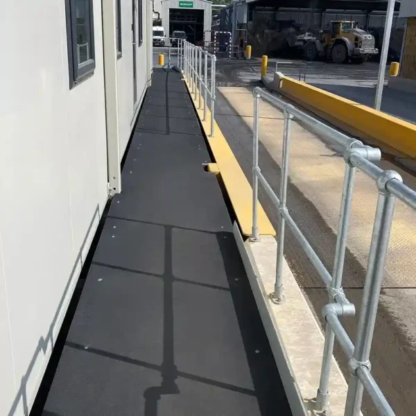 A black Anti-Slip Flat Sheet walkway with metal railings runs next to a white building, with industrial vehicles and equipment visible in the background beyond a yellow kerb.