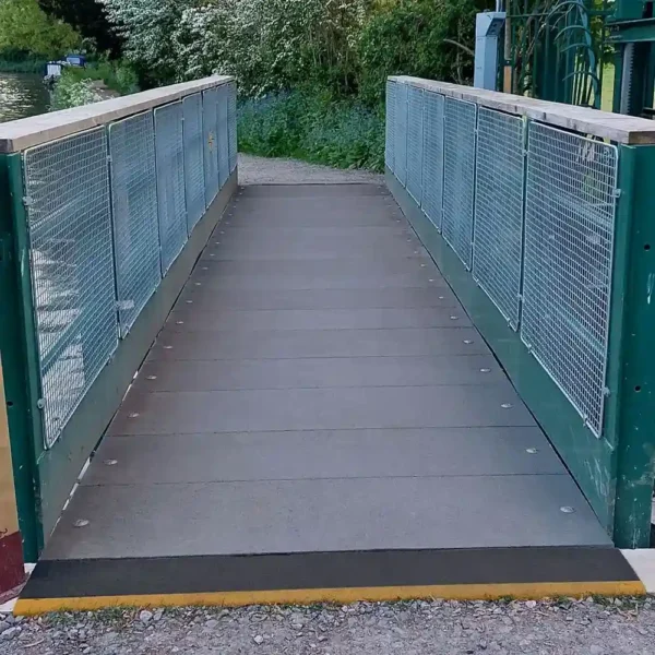A small footbridge with metal railings features an Anti-Slip Flat Sheet as its grey surface, connecting two gravel paths and surrounded by greenery and trees.
