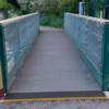 A small pedestrian bridge featuring Anti-Slip Flat Sheet decking, with green railings and metal mesh sides, spans a gravel path surrounded by greenery.