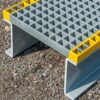A close-up of the Step Over Platform shows its grey metal grid surface with yellow edges on asphalt, highlighting its sturdy, industrial design for outdoor or heavy-duty use.