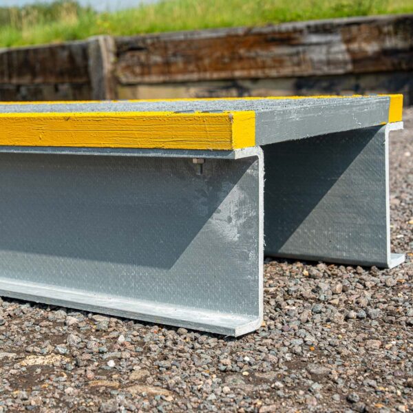 A close-up of the Step Over Platform shows its grey metal beam with a yellow stripe on top, resting on gravel, while grass and wooden planks are blurred in the background.