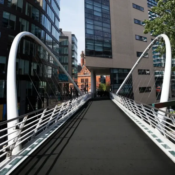 A modern pedestrian bridge with white railings and cables, equipped with the Anti-Slip Sheet, spans between office buildings and leads to a distant brick building with a clock tower.