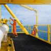 Wearing an orange boiler suit and helmet, a worker operates a valve on a yellow offshore oil platform featuring the Anti-Slip Sheet, with sea views and a lifebuoy hanging on the nearby railing.