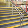 Indoor stairs with grey tiles feature Anti-Slip Stair Nosings in yellow, along with metal handrails and red railings, set against a brick wall background.