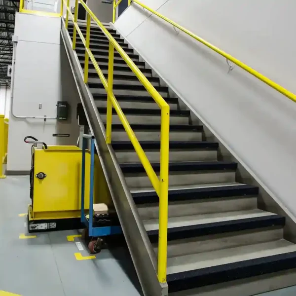 A grey staircase fitted with Anti-Slip Stair Nosing and yellow handrails rises beside a white wall. At the bottom, a yellow industrial trolley and blue platform trolley rest on a grey floor marked with yellow lines.