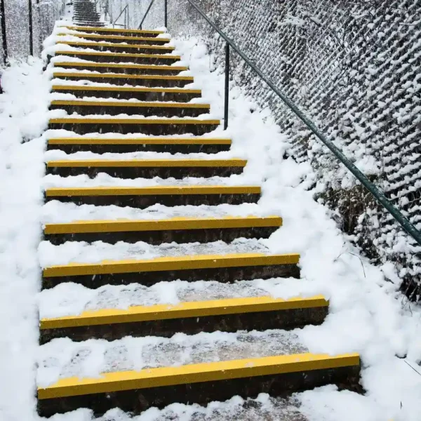 An outdoor staircase with Anti-Slip Stair Tread in yellow on each snow-covered step, bordered by a metal fence and handrail, on a winter’s day.