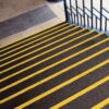 The Anti-Slip Stair Tread features dark steps with bright yellow safety lines on each edge beside a blue metal handrail, leading down to a tiled floor with a textured mat at the bottom.