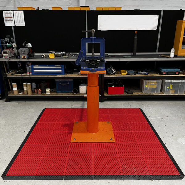 A blue and orange metal tool on an orange plinth stands on a red Multi-Purpose Interlocking PVC Tile mat in a workshop, with shelves of tools and equipment in the background.