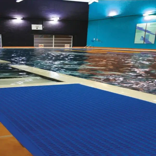 Indoor swimming pool with clear water, featuring a Leisure Mat in blue on the poolside. The area has blue and dark walls, bright ceiling lights, and a colourful artwork displayed on the right wall.