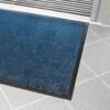 The Plush Entrance Mat, a blue rectangular doormat with a black rubber border, is placed on a light grey tiled floor near an entrance door with metal grooves.