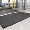 A Plush Entrance Mat in dark grey rests on a light grey tiled floor before a partially open glass entrance door, with cars visible outside through the glass.