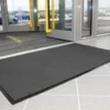 A Plush Entrance Mat in large black size is placed on a tiled floor by a glass door at a building entrance, with cars and trees visible outside through the windows.