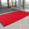 A plush entrance mat in bright red lies on a tiled floor inside a building near an open glass door with yellow handles, with cars visible outside through the glass.