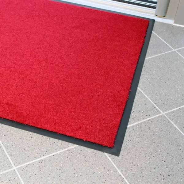 The Plush Entrance Mat, a vibrant red rectangle with a black border, is placed on a light grey tiled floor near a glass door.