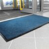 A Plush Entrance Mat is placed on a tiled floor by the building's glass doors with yellow handles, whilst cars can be seen outside.