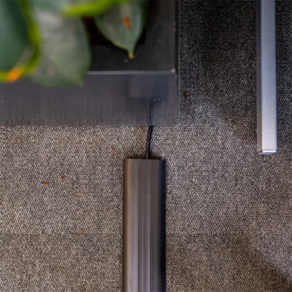 A black cable runs under a PVC Cable Protector on a grey carpeted floor, next to a black table with green leaves partially visible on top.