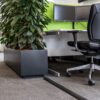 An office workspace with a black chair, a desk with dual monitors, a large rectangular planter holding a leafy green plant, and a PVC Cable Protector on the grey carpeted floor.