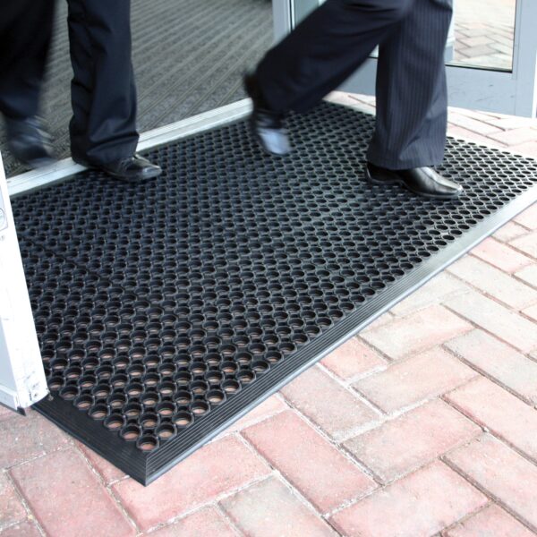 Two people in dark trousers and dress shoes walk across a Drainage Top Mat with circular holes, placed at a building entrance with glass doors and brick paving outside.
