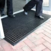 Two people in dress trousers and shoes step onto a Drainage Top Mat, a black rubber doormat with circular holes, placed at the entrance of a building with brick paving outside.