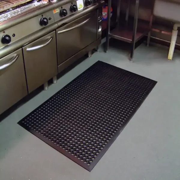 A black Drainage Top Mat with holes is placed on the grey floor in front of stainless steel kitchen appliances and a metal prep table in a commercial kitchen.