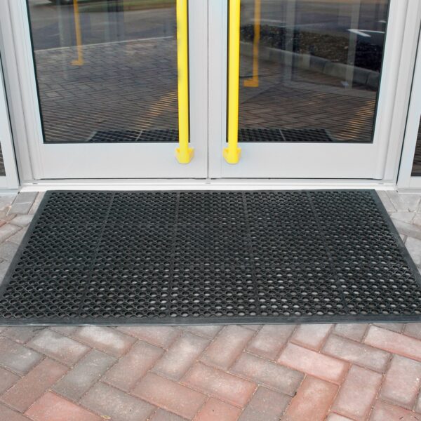 The Large Front Door Mat Bundle, a black rubber mat with holes, is set before a glass double door with yellow handles on interlocking brick paving.