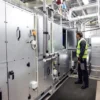 Wearing safety gear and a high-visibility vest, a person inspects large industrial HVAC equipment inside a facility with metal pipes and ducts, whilst standing on Electrical Safety Matting for added protection.