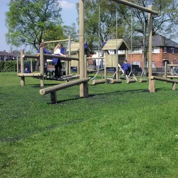 Children play on grass protected by the 16mm Black Grass Mat, climbing a wooden frame and swinging in a sunny playground bordered by trees and houses.