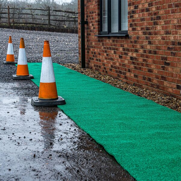 Three orange and white traffic cones are set on a wet surface beside a Heavy-Duty PVC Vinyl Roll, placed along a brick building, with gravelly ground and a wooden fence visible in the background.