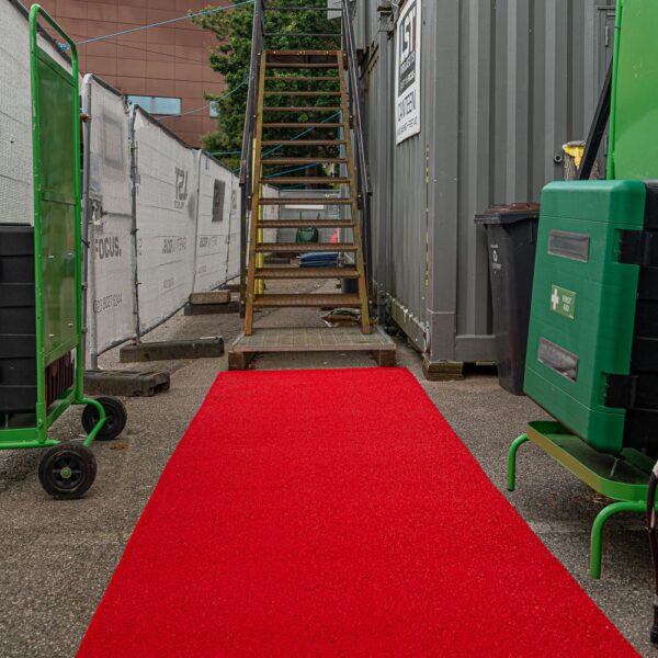 A Heavy-Duty PVC Vinyl Roll in bright red leads to a metal outdoor staircase, surrounded by construction barriers, green trolleys, and equipment cases for an industrial, utilitarian setting.