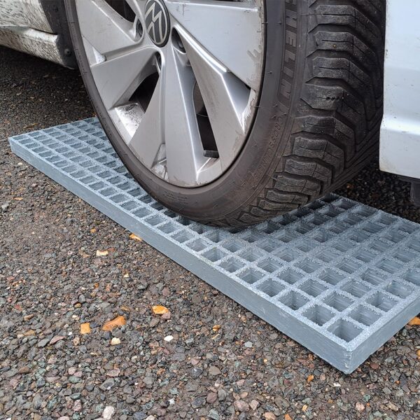 A car tyre rests on Waffle Boards, rectangular metal grates, which are placed on gravel beside a white vehicle.